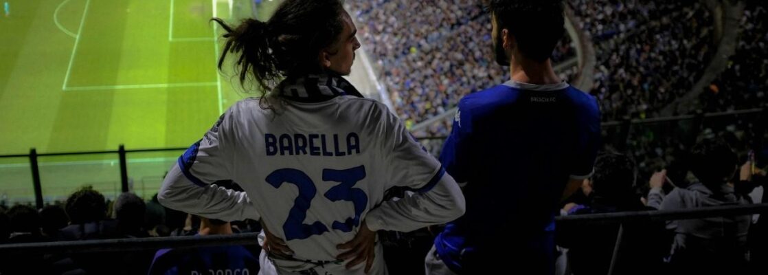 A behind view of soccer fan wearing number 23 jersey, with his hands on his waist looking at another fan, both facing the football pitch from the fans' stand.