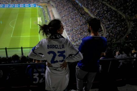 A behind view of soccer fan wearing number 23 jersey, with his hands on his waist looking at another fan, both facing the football pitch from the fans' stand.